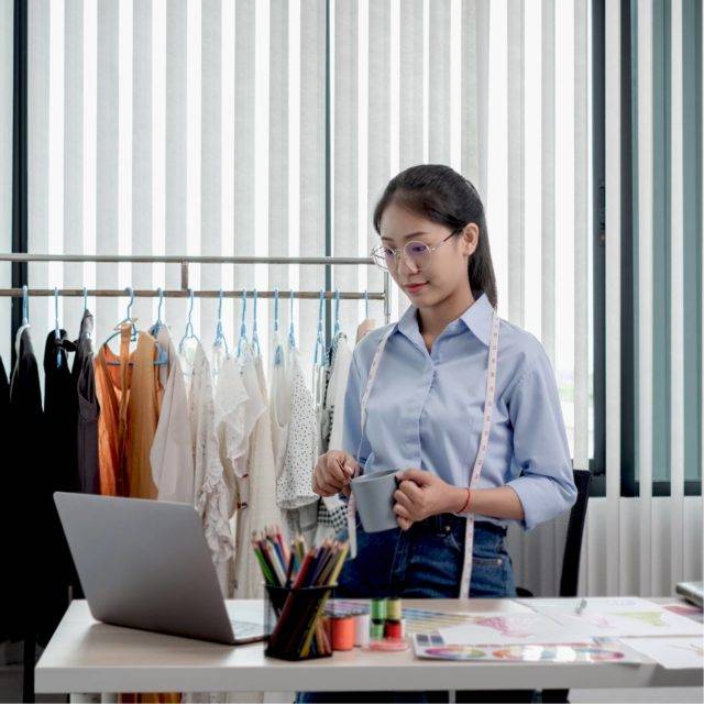 Women standing up at desk in front of laptop with a rail of clothes behind