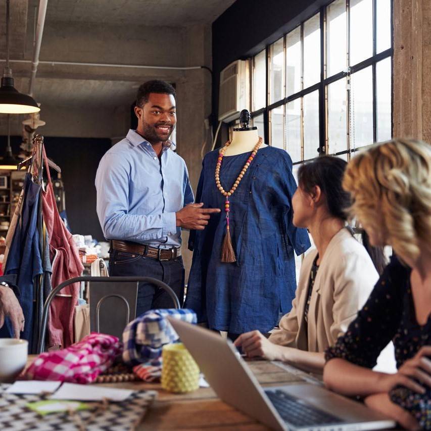 Male fashion designer presenting blue garment to two women in factory