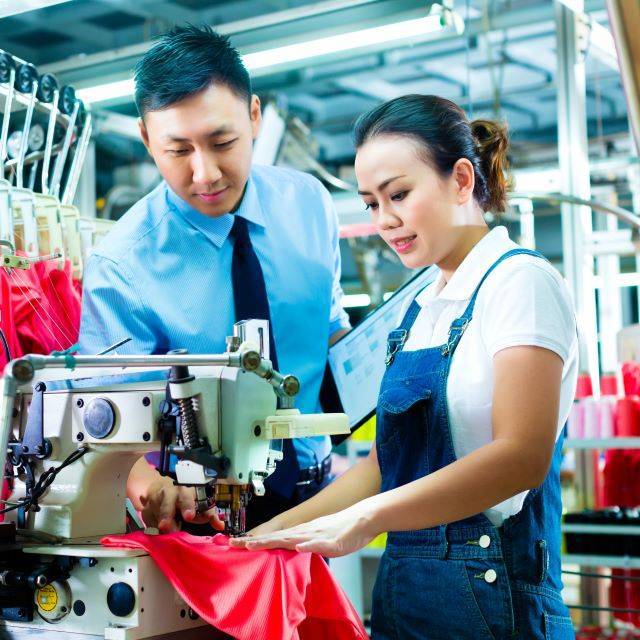 Man wearing tie observing female seamstress wearing dungarees using a sewing machine in garment factory